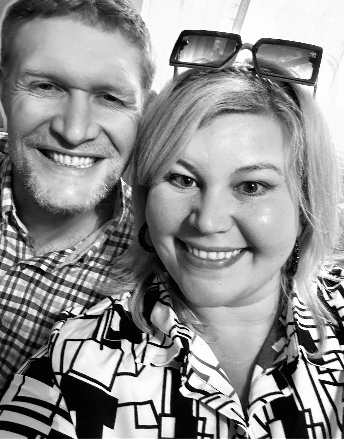 A black and white close-up photograph of a smiling man and woman, both wearing patterned clothing. The woman has sunglasses resting on her head.