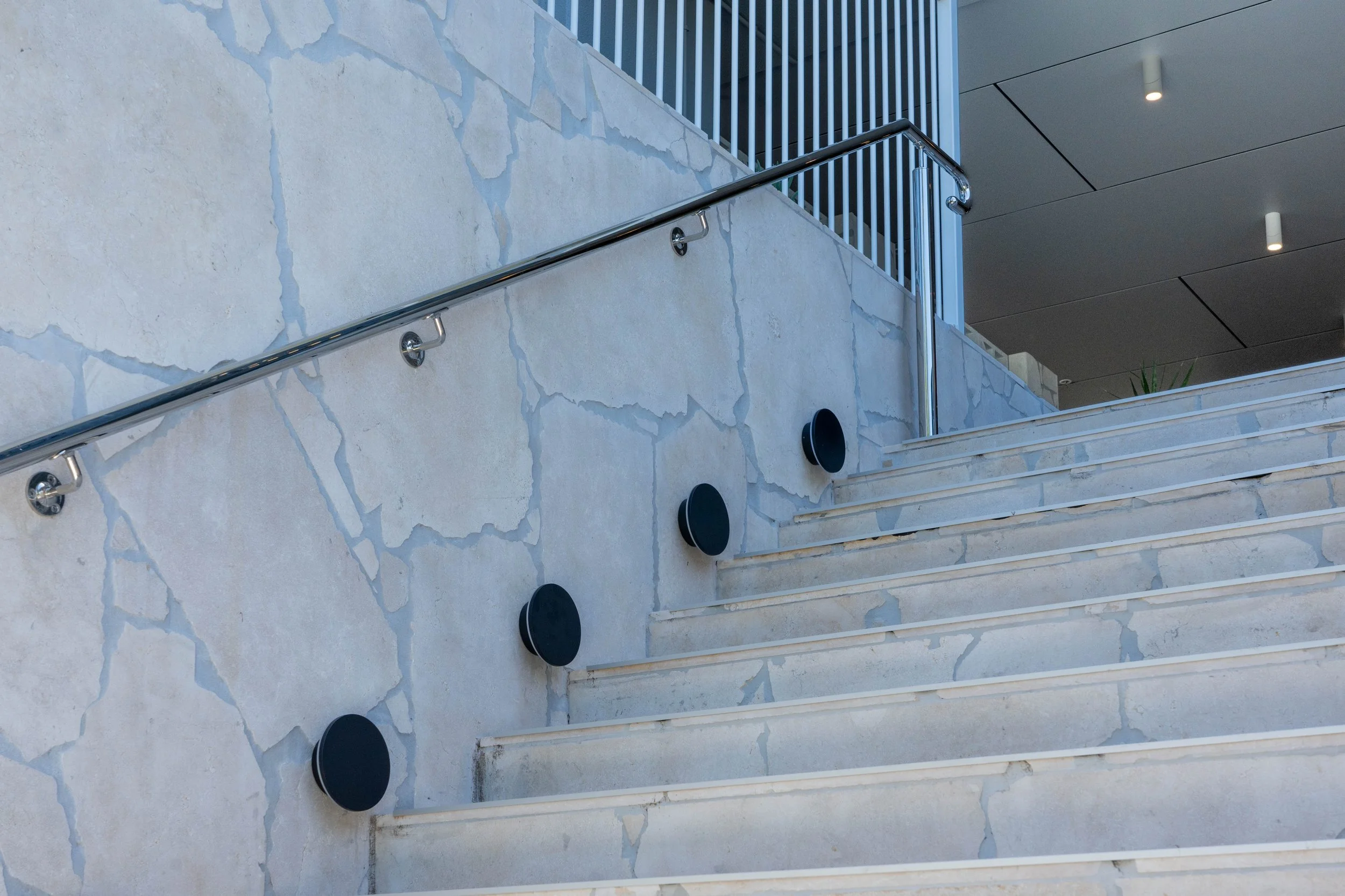 Outdoor staircase with a metal handrail, black circular sensors or connectors on the wall, and a modern building ceiling with recessed lighting.