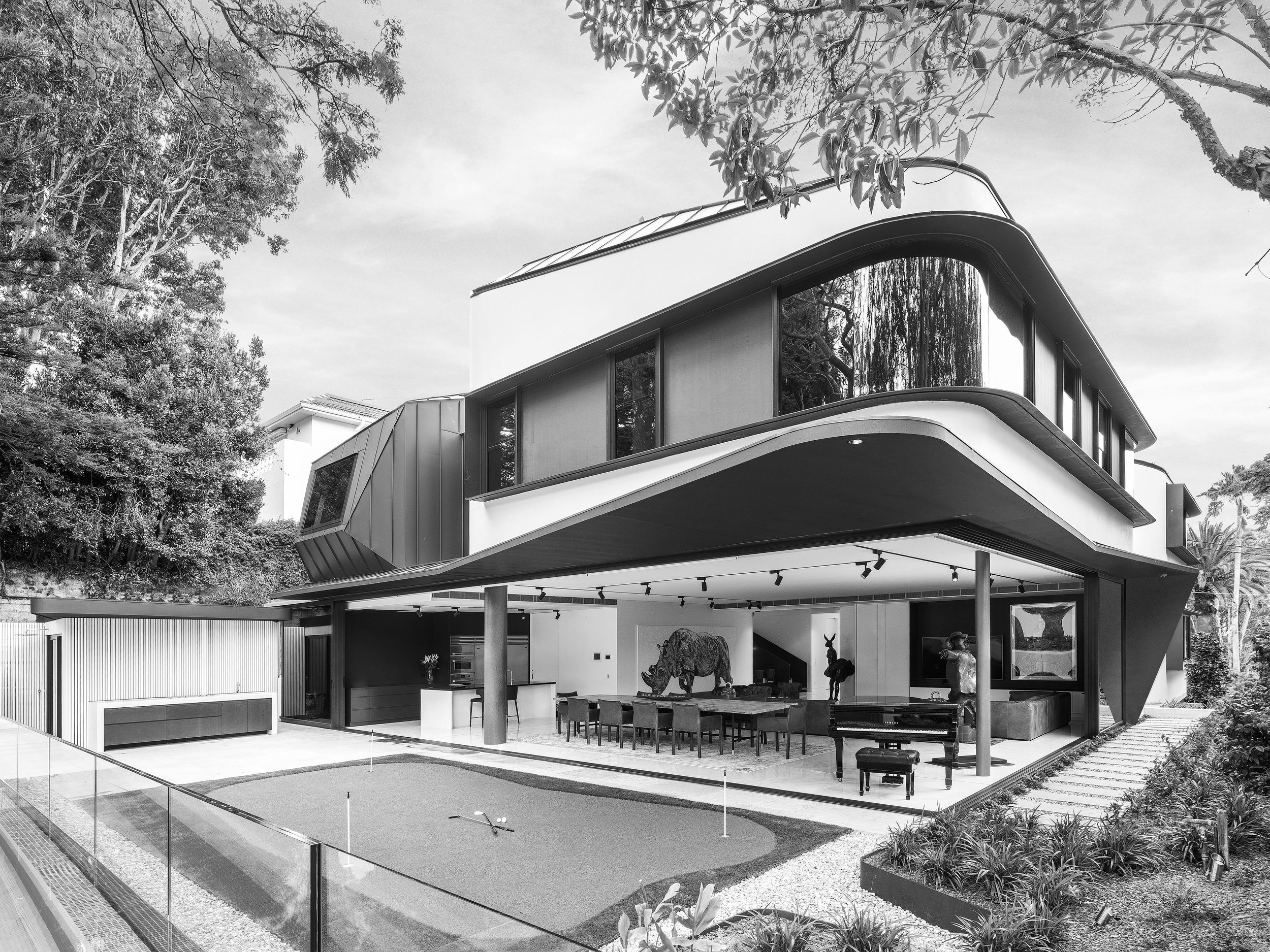 A modern house with large windows and a balcony, surrounded by trees, with an outdoor area that includes a putting green and a seating area, taken from a low angle in black and white.