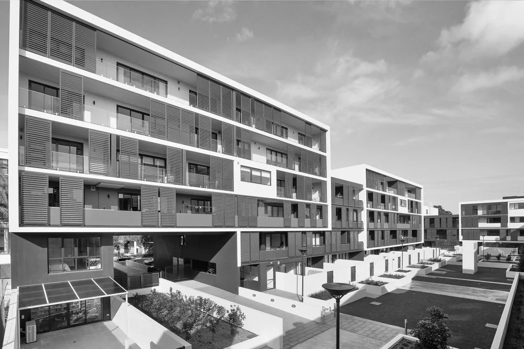 Black and white photograph of modern apartment buildings with multiple balconies and a landscaped courtyard with pathways and benches.