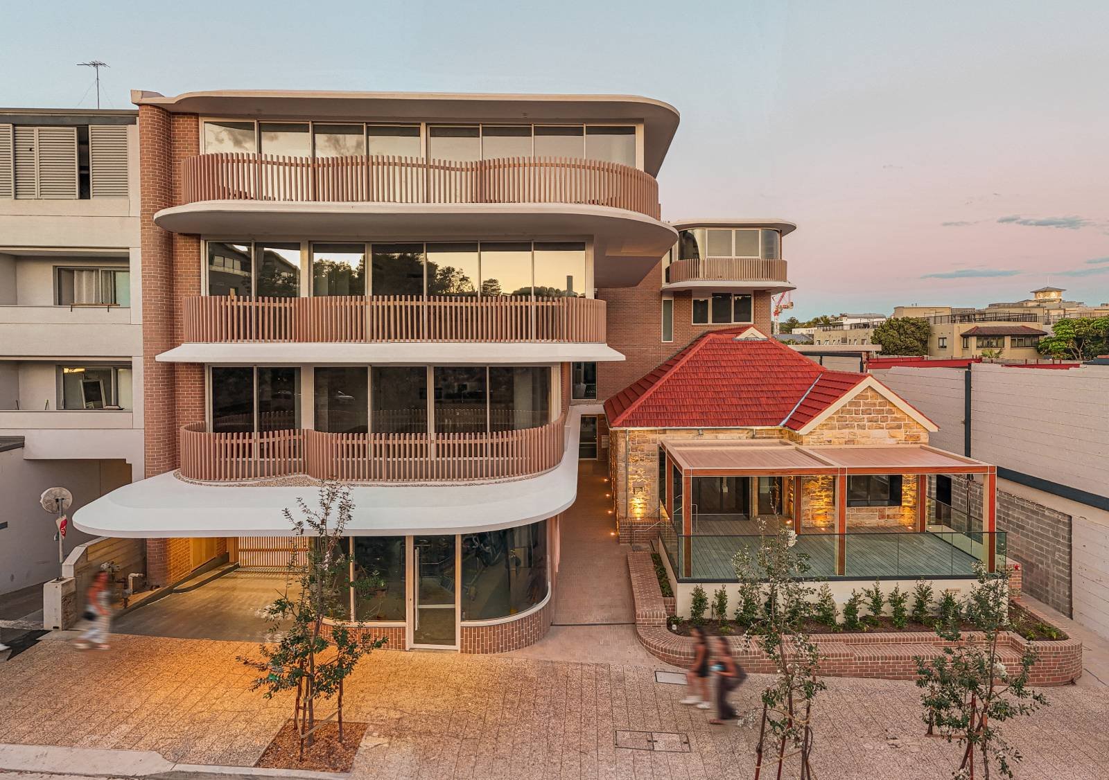 Multi-story modern building with curved balconies, a brick tower, and a glass-enclosed terrace, set against a sunset sky in an urban area.