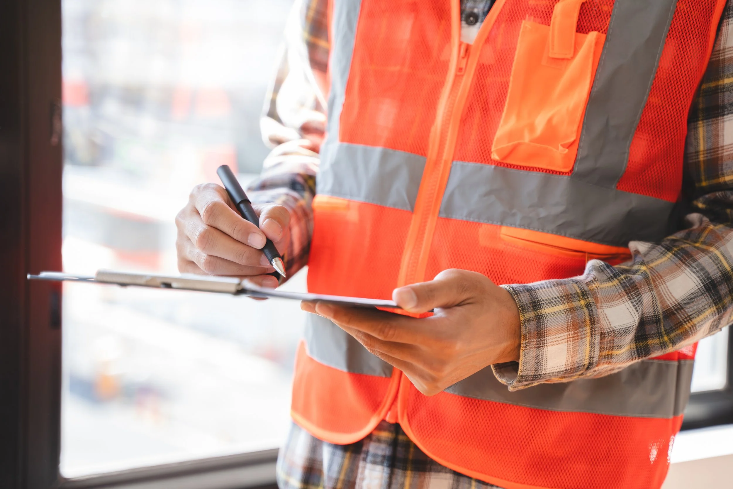 A construction worker in an orange safety vest writing on a clipboard with a pen.