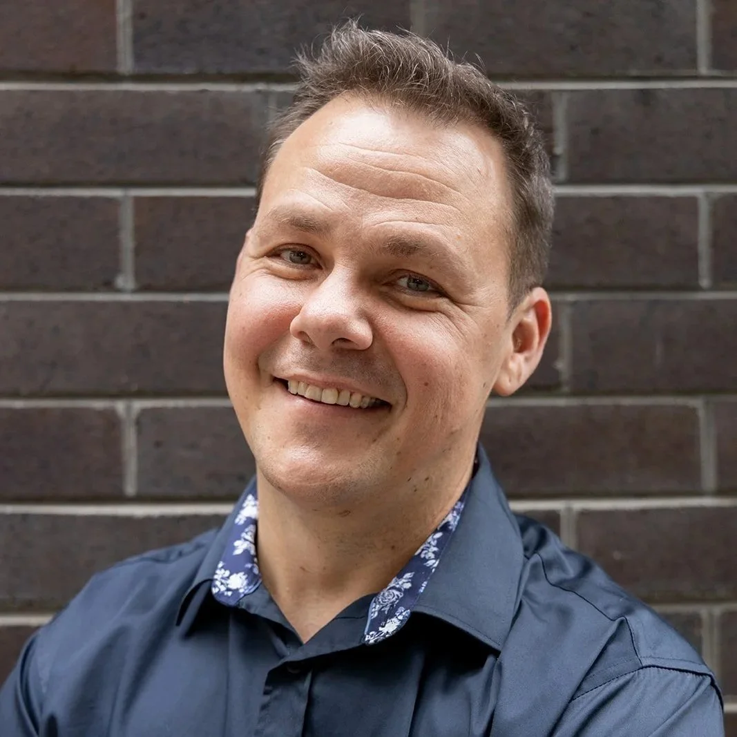 A smiling man with short brown hair wearing a dark blue shirt with a patterned collar, standing against a brick wall.