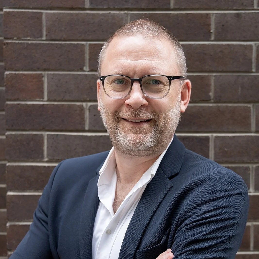 A smiling man with glasses, a beard, wearing a navy blazer and white shirt, standing in front of a brick wall.
