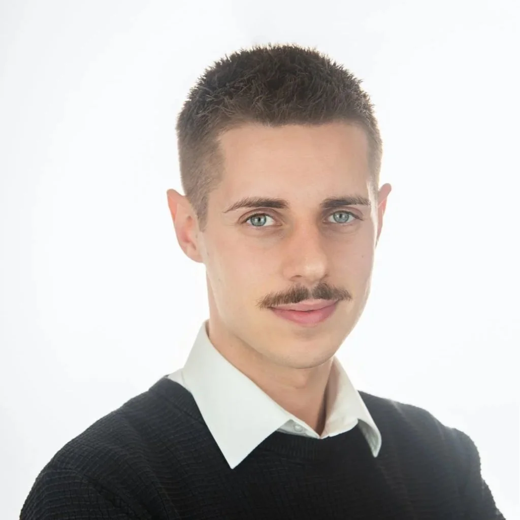 A man with short dark hair, a mustache, blue eyes, wearing a white collared shirt under a black sweater against a plain white background.
