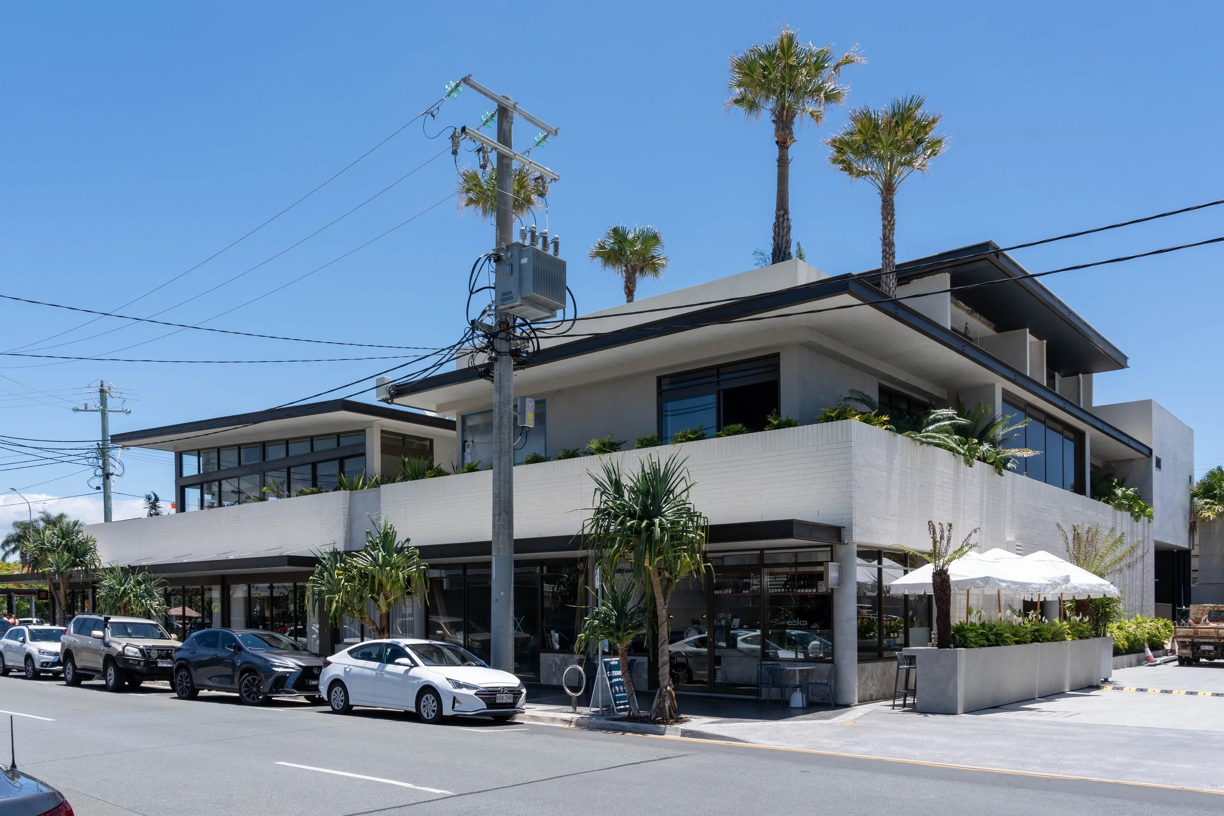 Modern multi-story building with large glass windows, surrounded by tall palm trees and parked cars in a sunny urban area.