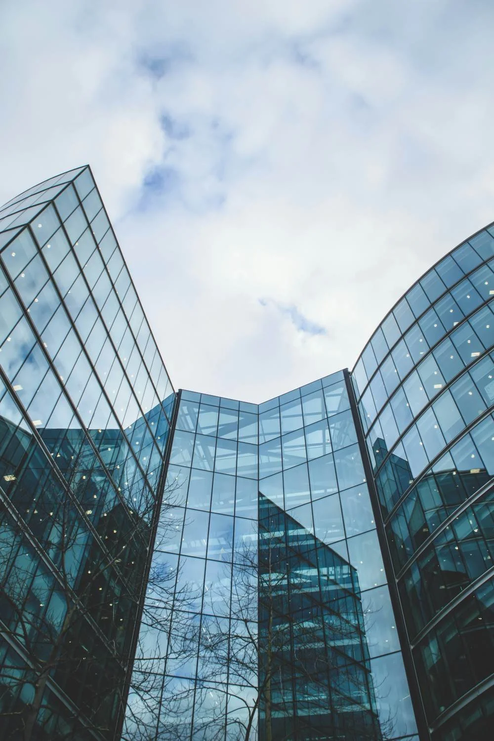 Low-angle view of modern glass office building with reflective windows against a cloudy sky.