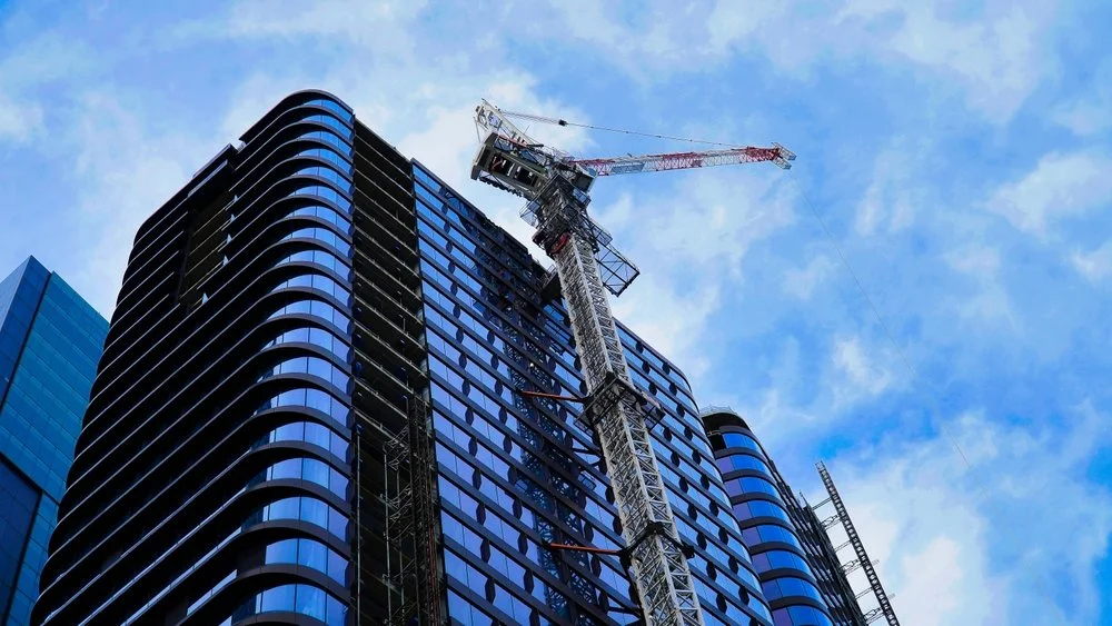 A high-rise building under construction with a crane on top against a blue sky with clouds.