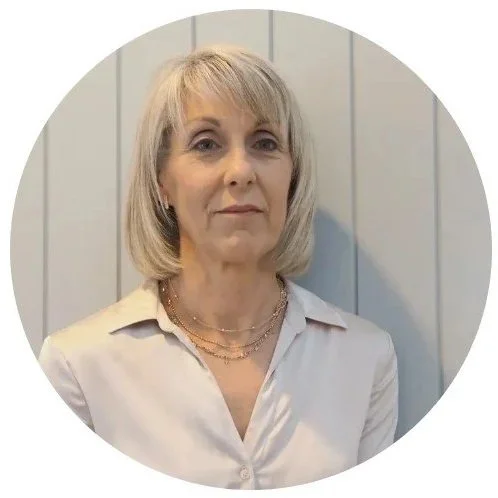 A middle-aged woman with shoulder-length blonde hair standing against a light gray paneled wall, wearing a light-colored blouse and layered necklaces.
