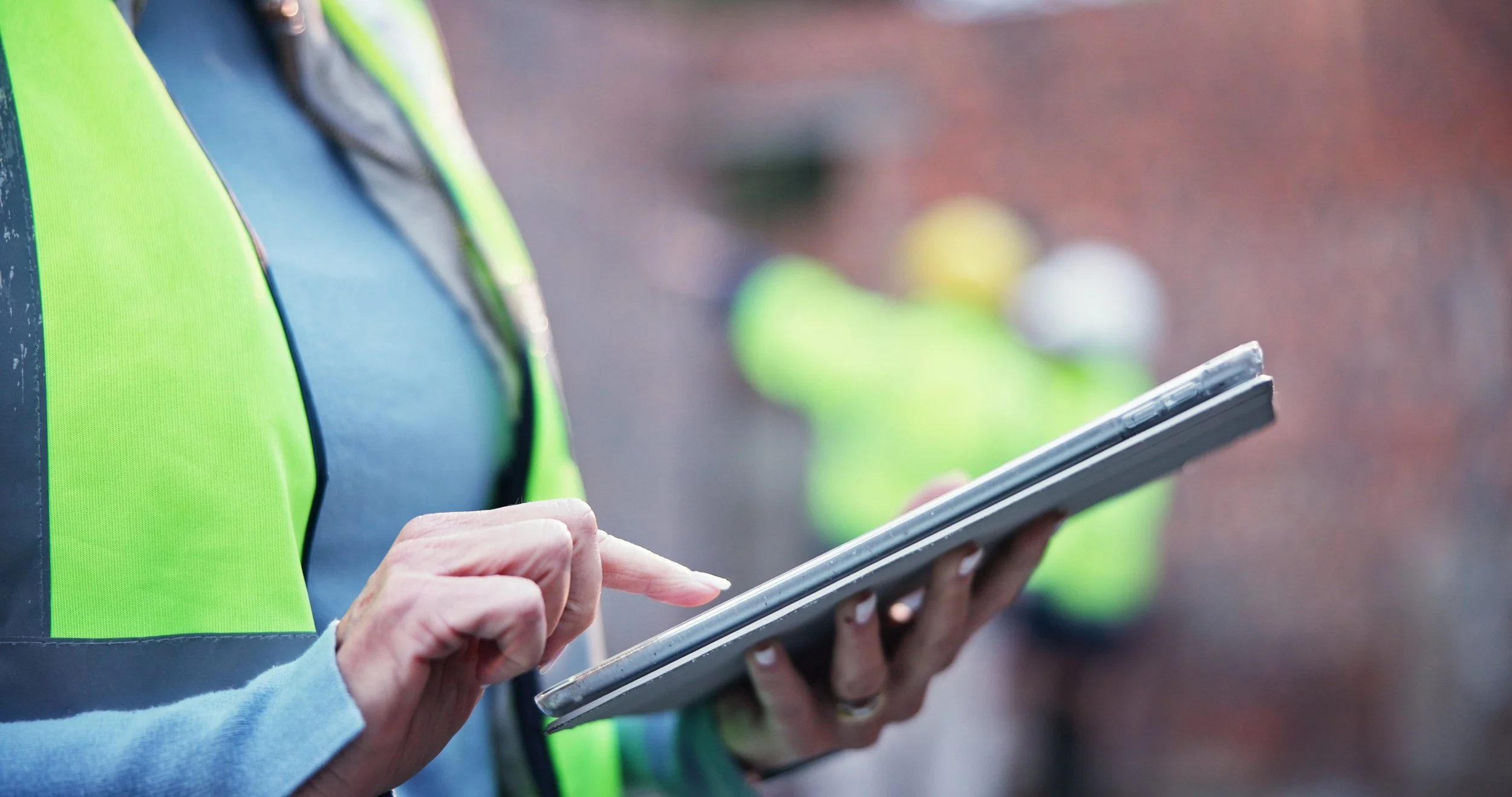 Person in a high visibility vest holding and using a tablet outdoors, with other people in high visibility vests blurred in the background.