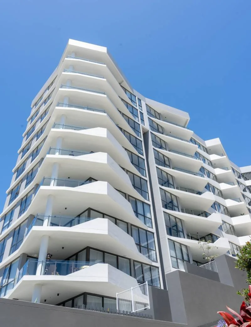 Modern multi-story apartment building with white balconies and glass railings against a clear blue sky.