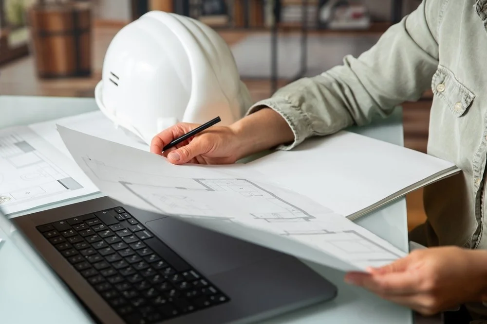 A person reviewing architectural blueprints at a desk with a laptop and a white safety helmet in the background.