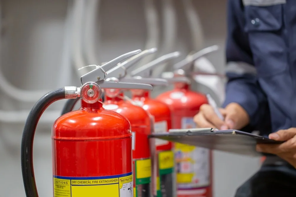 Several red chemical fire extinguishers lined up in a row with a person in a dark blue uniform writing on a clipboard nearby.