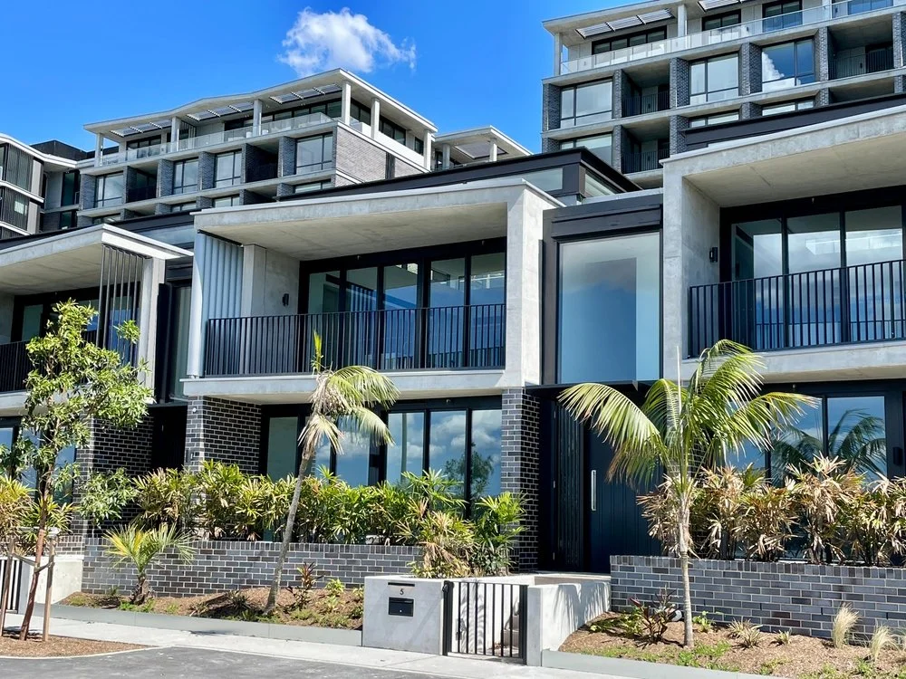 Modern multi-story residential buildings with large glass windows, balconies, and lush tropical landscaping in front.