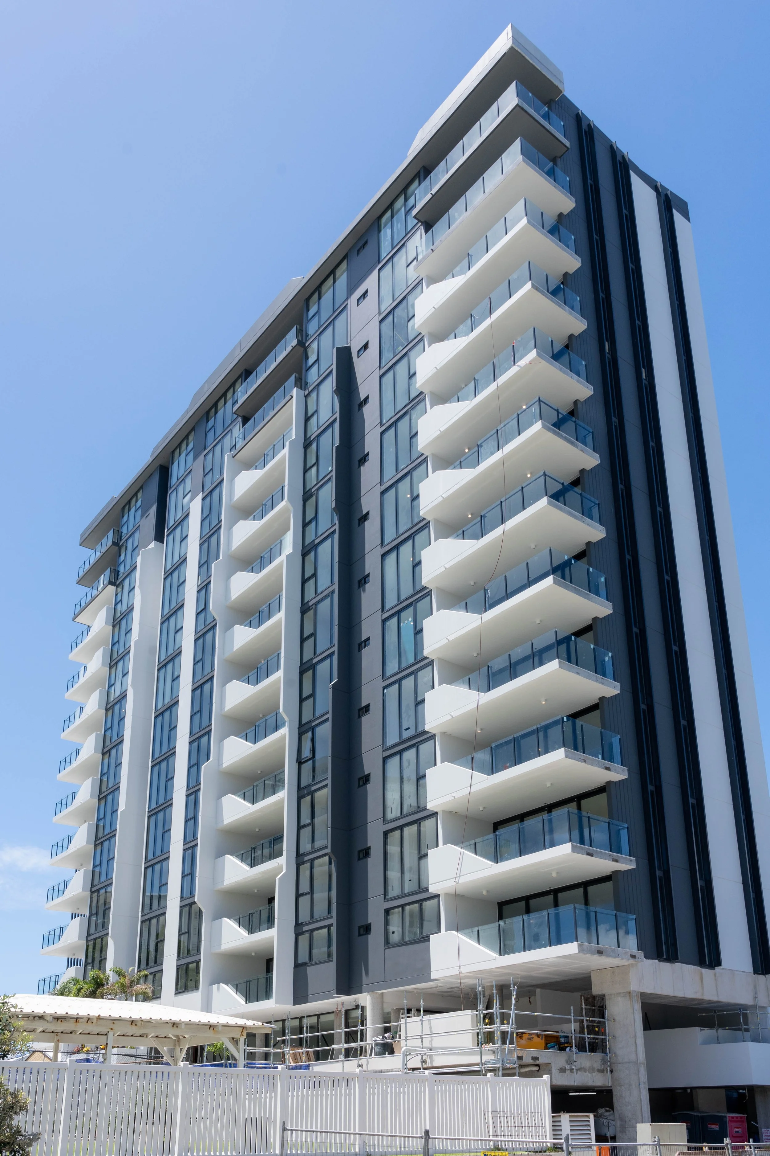 Modern multi-story apartment building with white balconies and glass railings, under construction, against a clear blue sky.