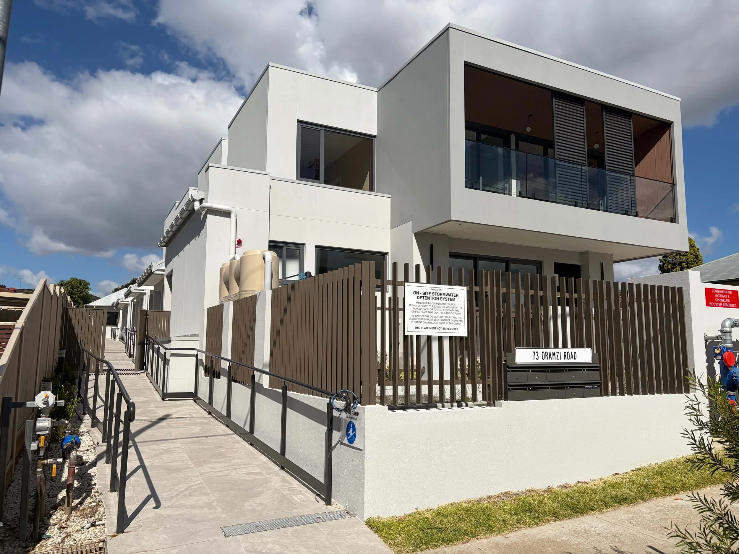 Modern white multi-story residential building with balcony and glass railing, situated under a cloudy sky. A ramp and pathway lead to the entrance, with plants and safety signs visible in the foreground.