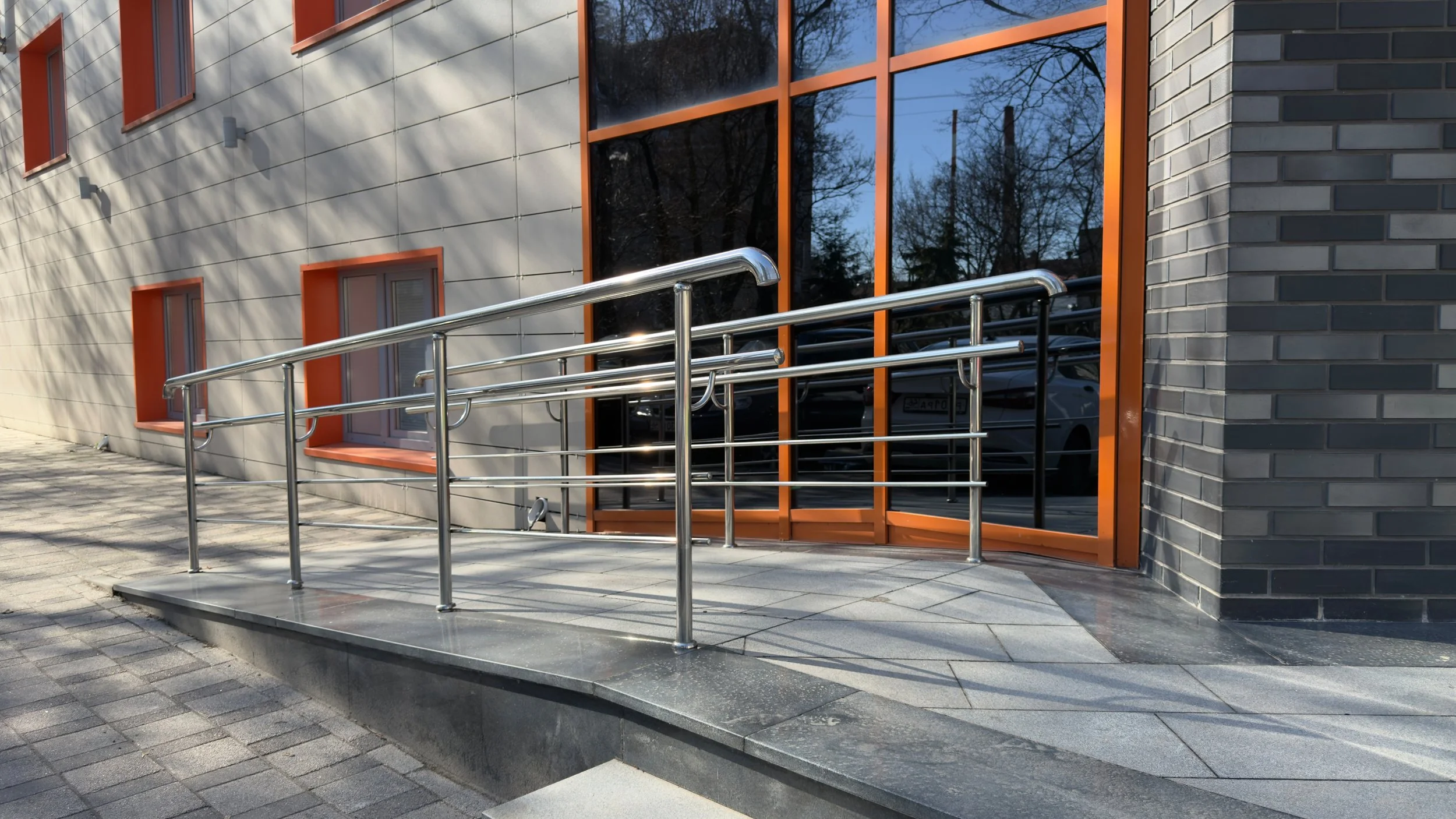 Ramped entrance with guard rails outside a modern building with gray brick and orange window frames.