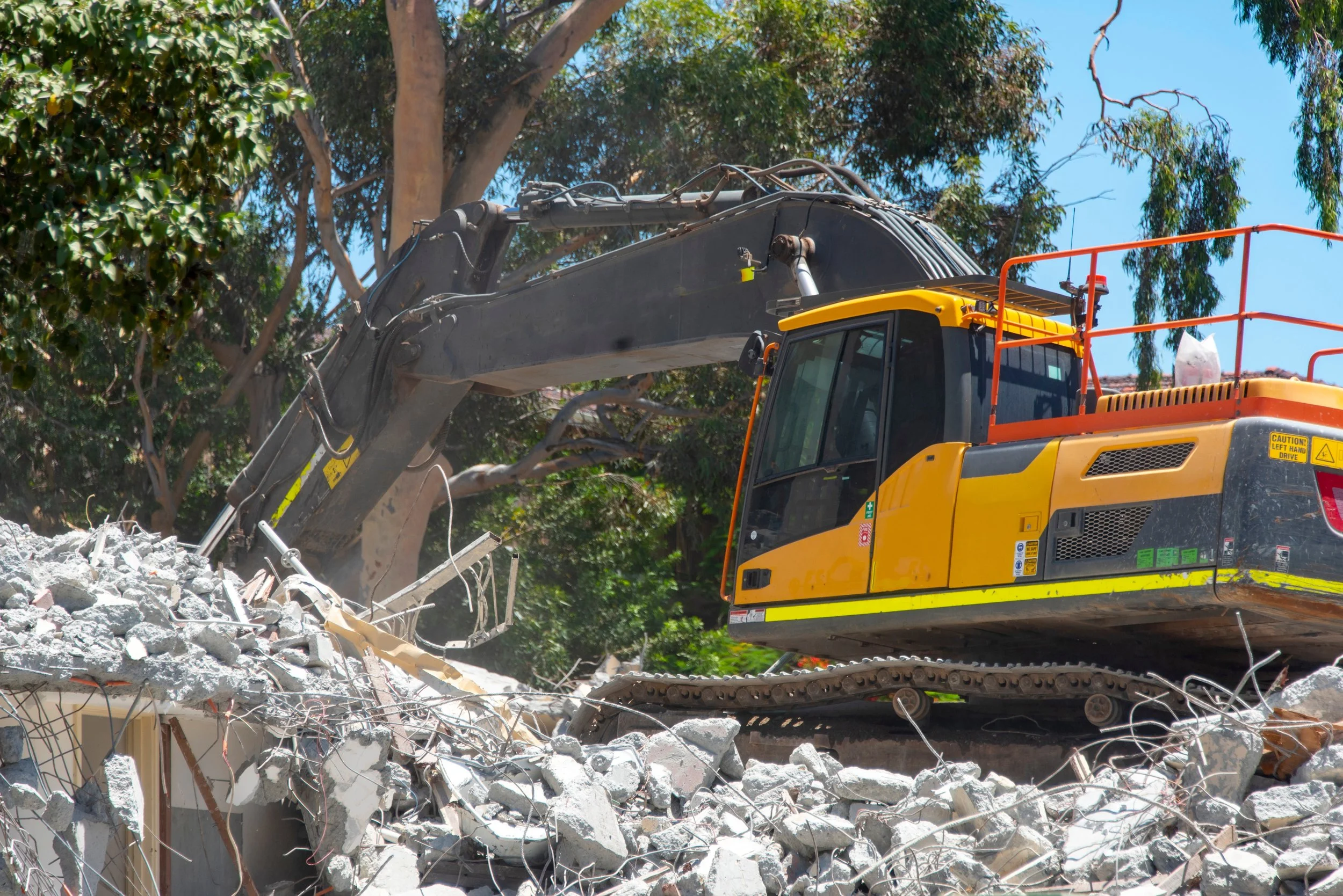 A yellow and black excavator crushing concrete debris at a demolition site, with trees and a blue sky in the background.