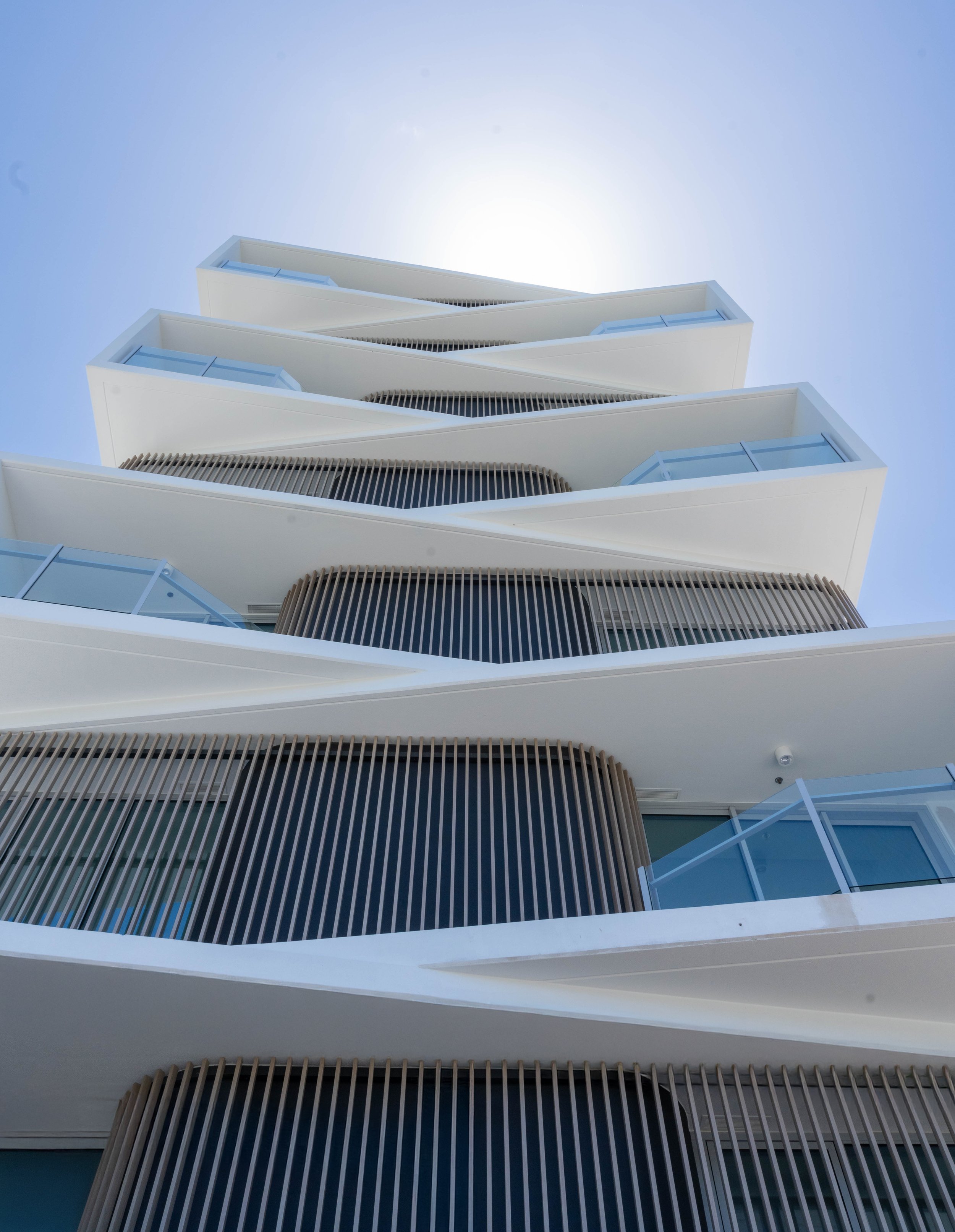 Low-angle view of a modern multi-story building with white balconies, glass railings, and vertical wooden slat design elements, with the sky in the background.