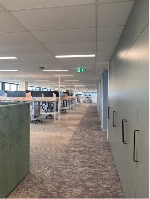 Empty office space with rows of desks, chairs, and computers, and a corridor with cabinets on the right.
