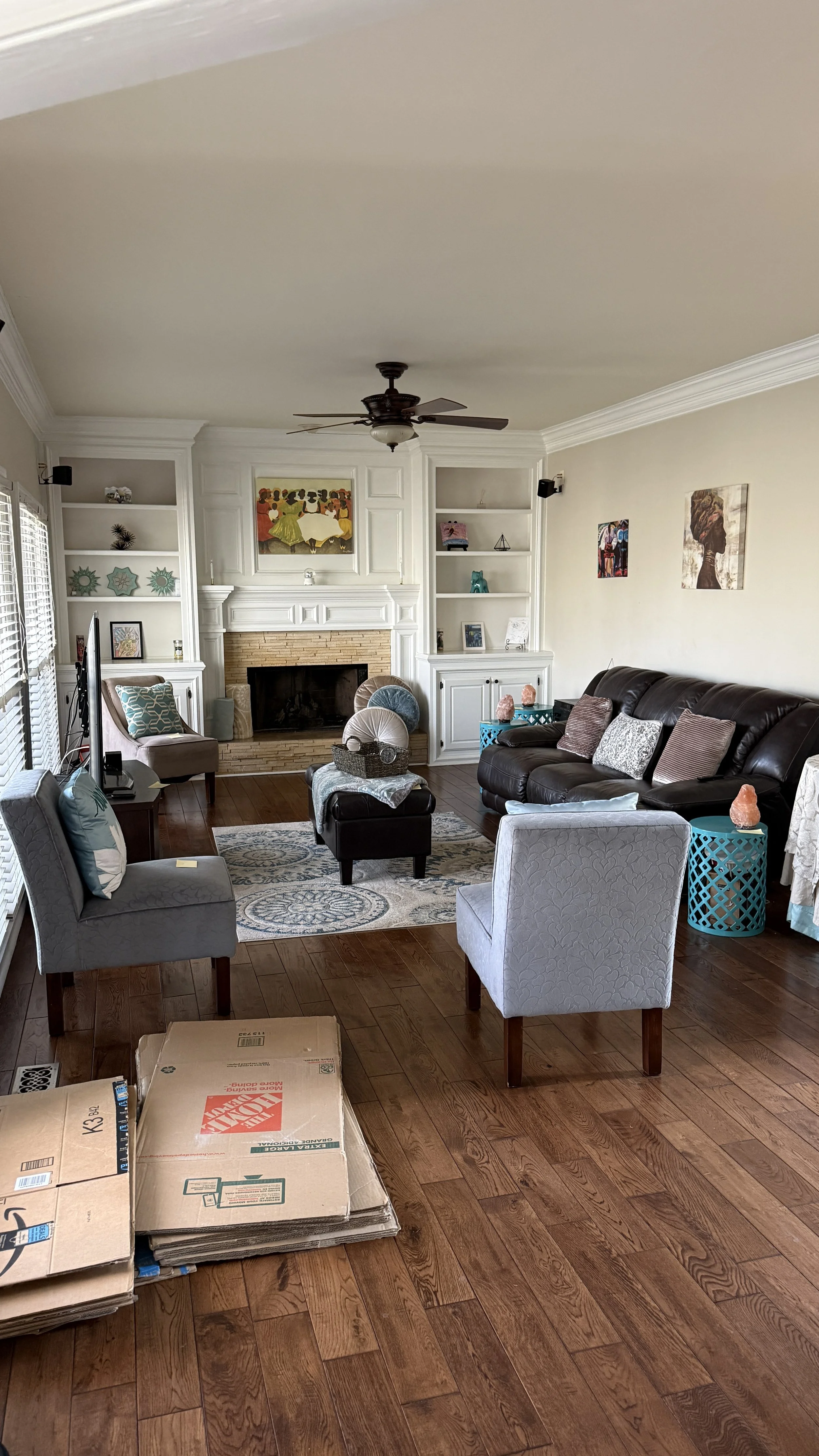 Living room with wood floors, white walls, built-in shelves, a fireplace, ceiling fan, and various seating including armchairs and a couch. There are decorative items on shelves, artwork on walls, and boxes on the floor.