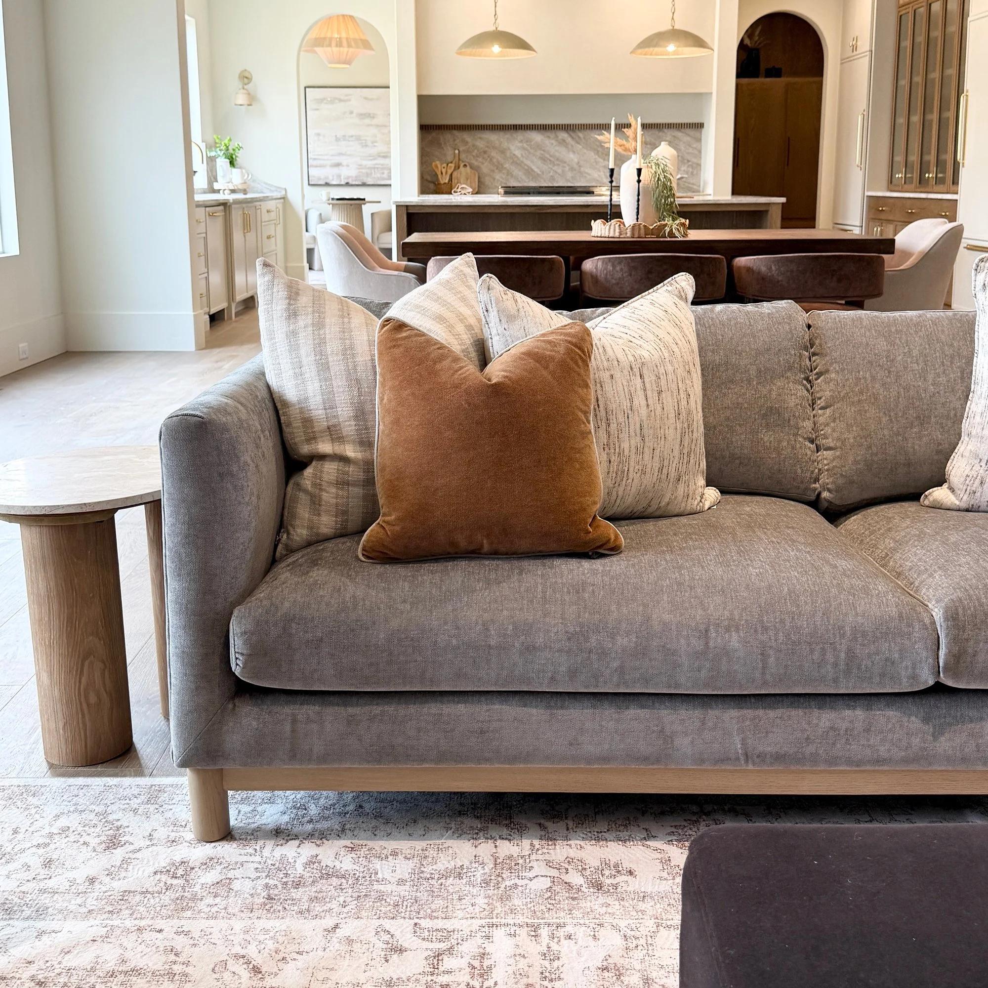 A modern living room with a sofa, pillows, and a carpet visible. In the background, there is a dining table with decorative vases and candles, along with a kitchen area and pendant lights hanging from the ceiling.
