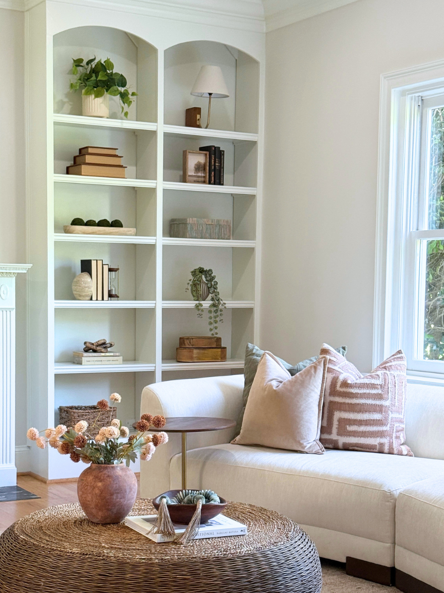 Bright, neutral living room featuring a cream sofa with blush and patterned accent pillows, white built-in arched bookshelves styled with books, greenery, and decorative objects, and a round woven cof