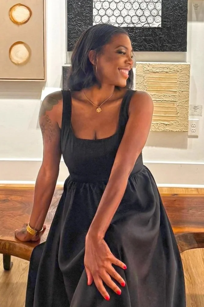 A woman in a black sleeveless dress, smiling and sitting on a wooden table in an art gallery or museum, with abstract textured art pieces hanging on the wall behind her.
