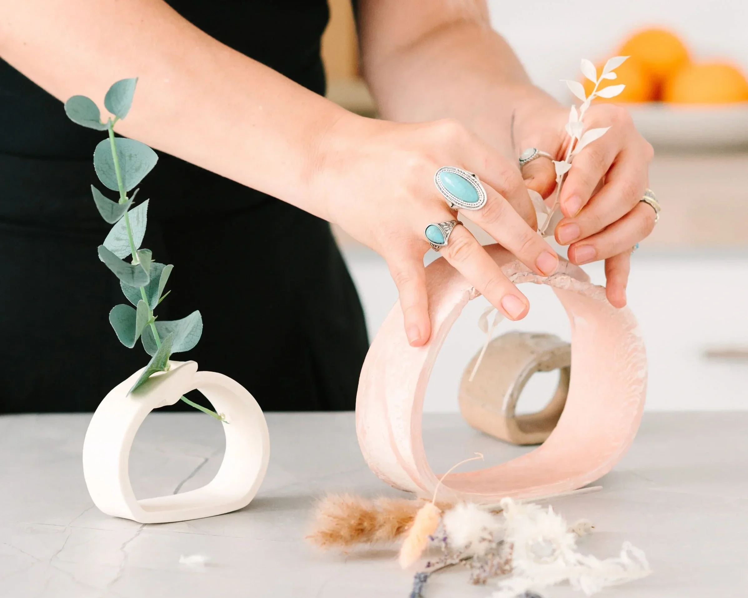 Close-up of a pink flamingo sculpture with a hollow body and long neck, with remnants of shredded cotton or stuffing on a flat surface in front of it. A green eucalyptus branch is also visible in the foreground, and person's hands are seen holding a 