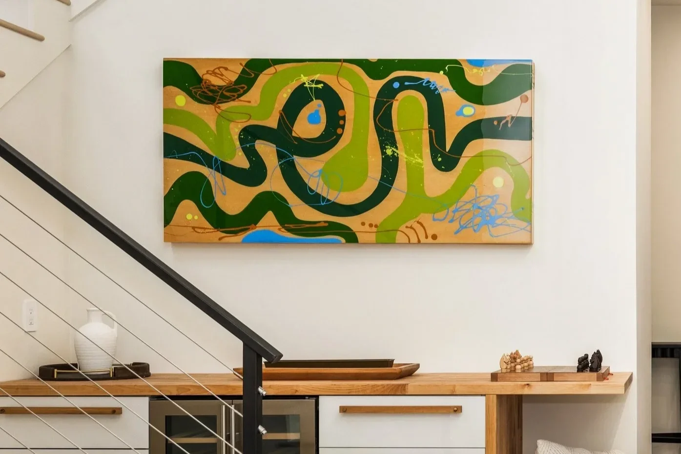 Colorful abstract painting with green, blue, black, and tan swirls and scribbles, hanging on a white wall in a modern home interior.