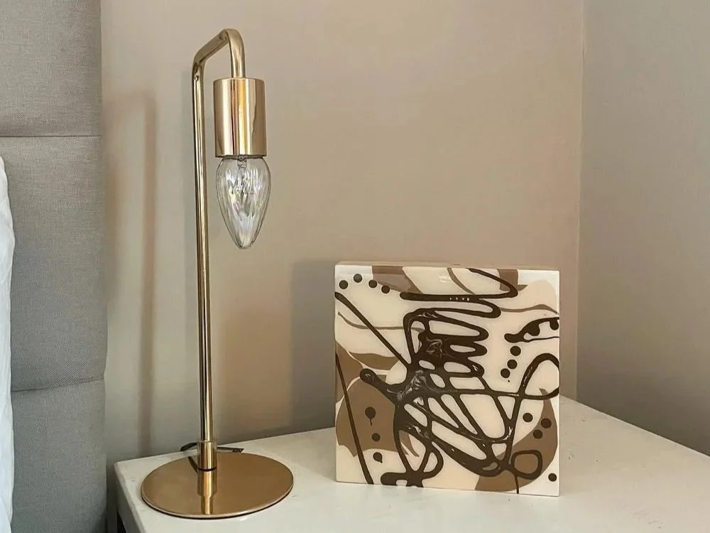 A bedside table with a gold-colored modern table lamp and an abstract painting in black, brown, and beige on a white canvas.
