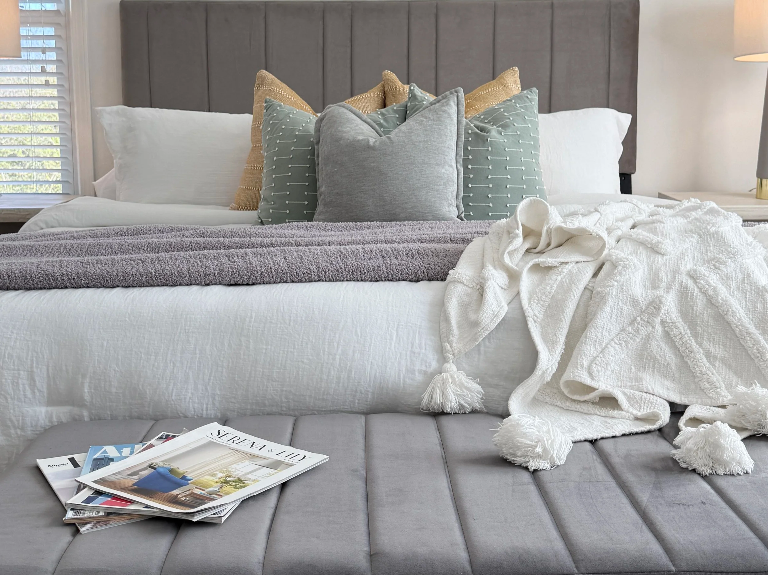 A bed with gray upholstered headboard, white sheets, and multiple decorative pillows in beige, gray, and light blue. A white textured blanket is draped over the bed. A stack of magazines or catalogs is on a gray upholstered bench at the foot of the b