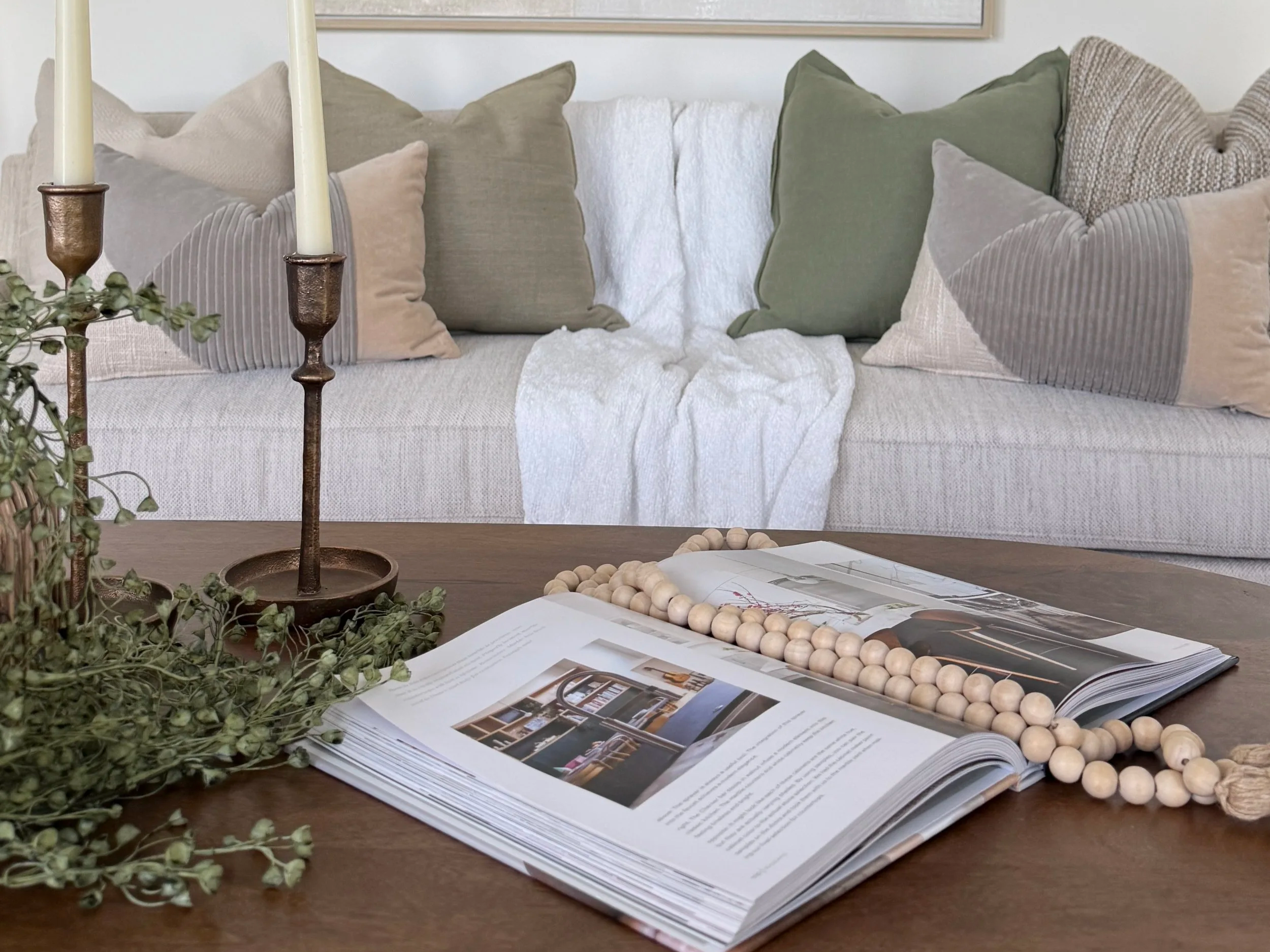 A wooden table with open magazines, a beaded necklace, and a candle holder with two candles, in front of a sofa with multiple beige, gray, and green pillows.