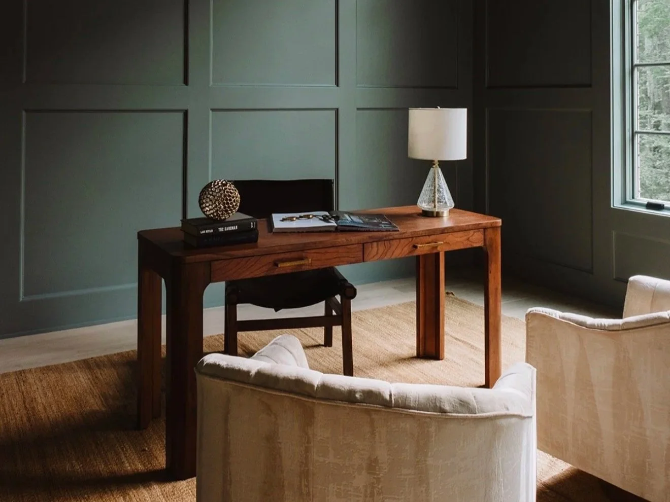 A cozy home office with a wooden desk, white armchairs, a lamp, a window, and decorative items on the desk, against paneled dark green walls.