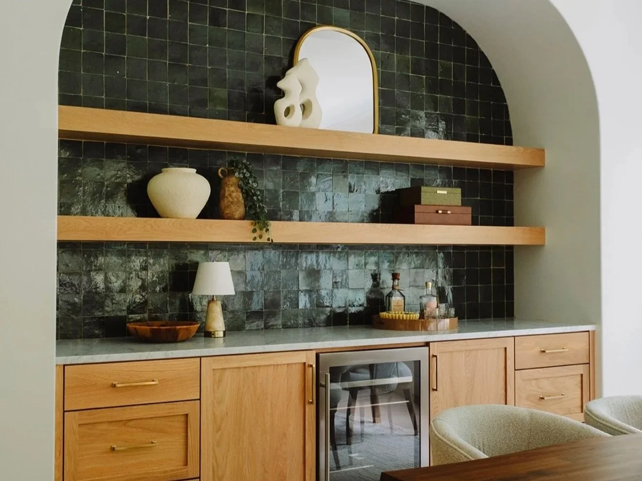 A built-in wooden bar area with closed cabinets, a small wine fridge, and an off-white countertop. Two open shelves hold decorative items, including vases, boxes, and a mirror. The black tiled wall behind features glossy square tiles.