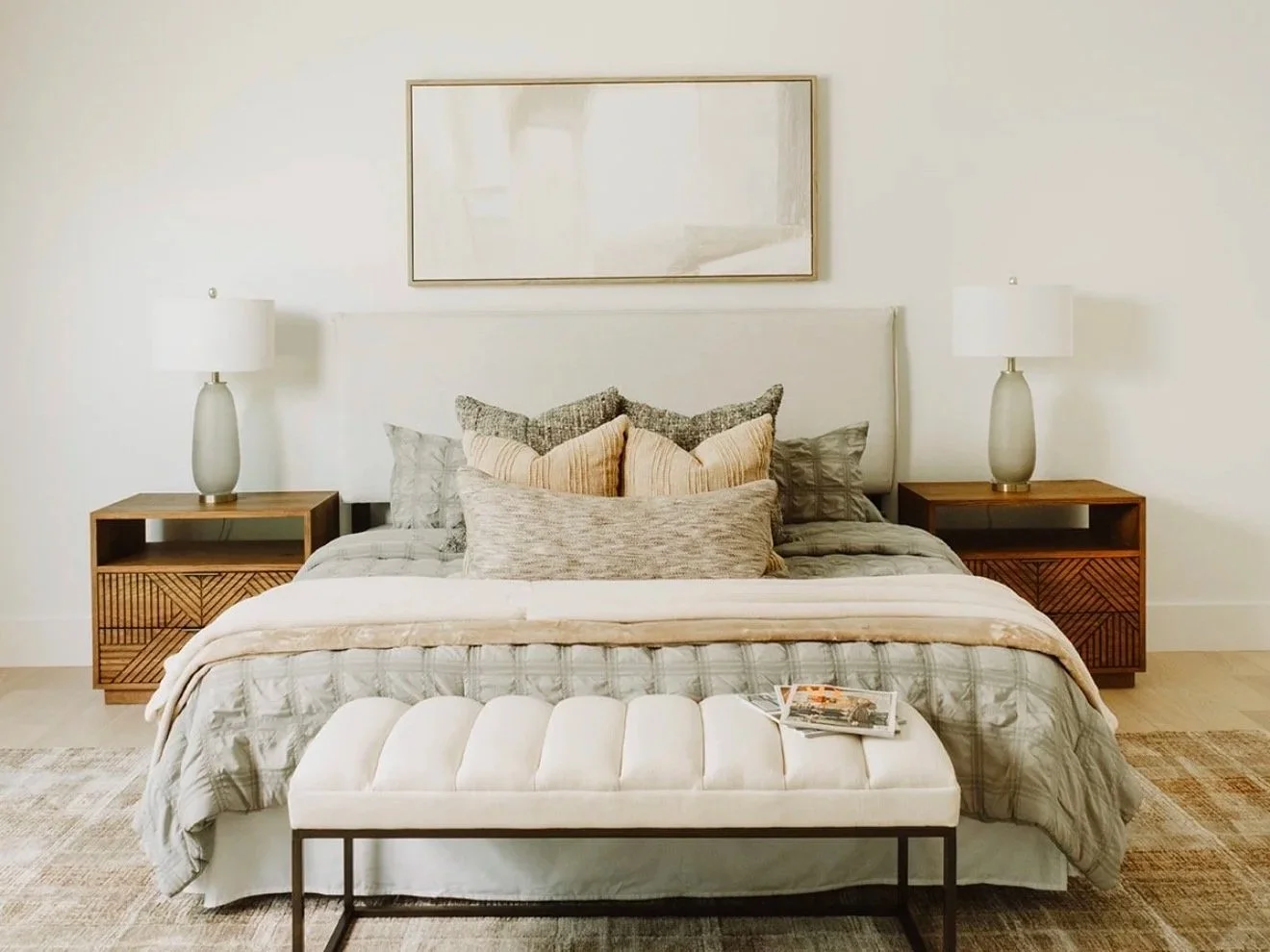 A neatly made bed with multiple pillows, flanked by two wooden nightstands with lamps, a large framed mirror above, and a white ottoman in front with a magazine on top, in a bright, minimalist bedroom.