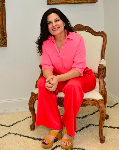 A woman with dark hair sitting on a vintage upholstered chair, smiling, wearing a pink blouse and coral-colored pants, with beige sandals, in a room with white walls and a rug.