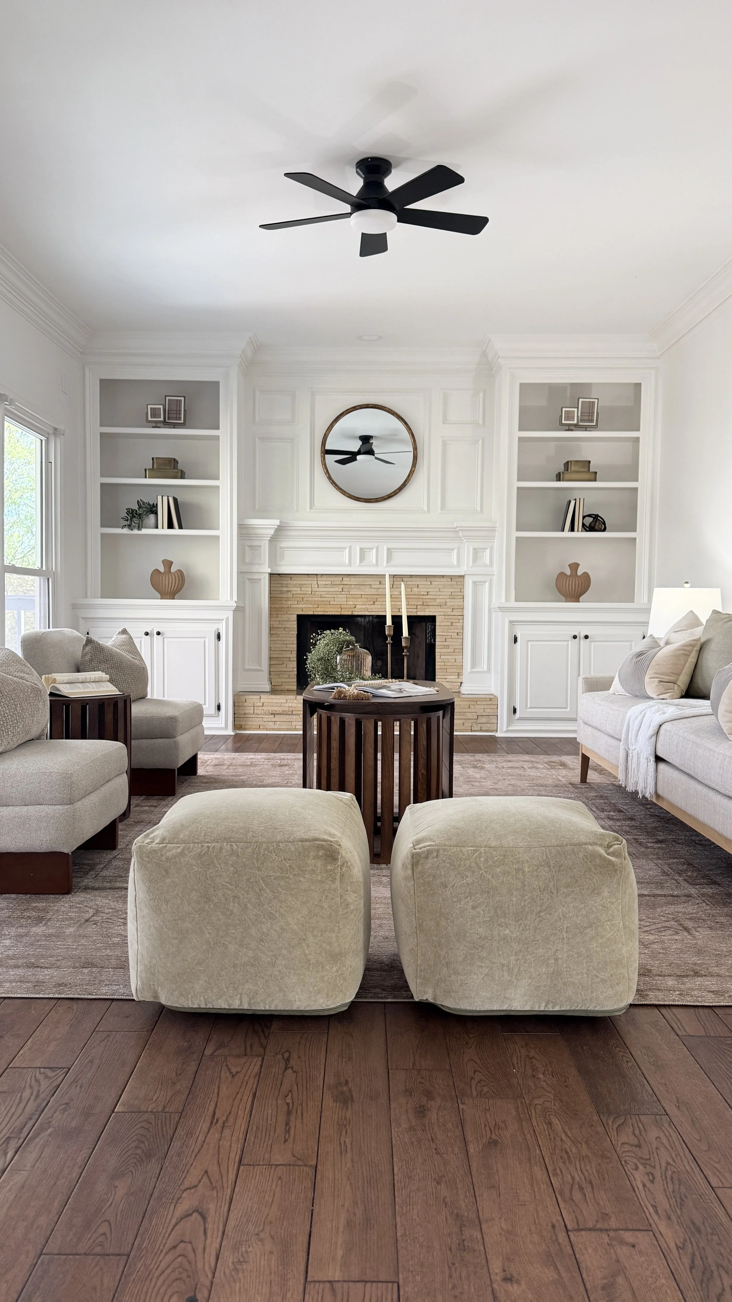 Living room with white walls, built-in shelves, a brick fireplace, beige sofas, a round wooden coffee table, a black ceiling fan, and a large window.