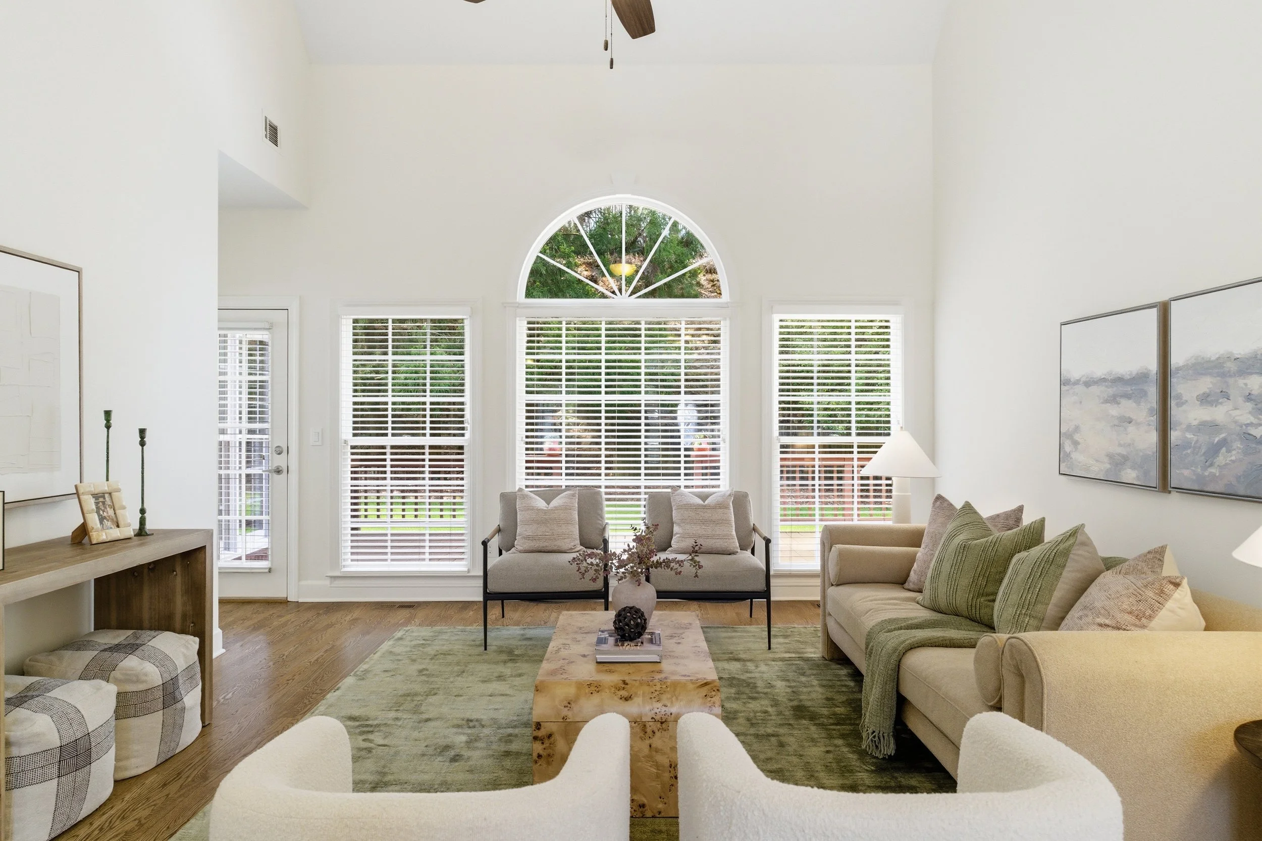 Bright living room with large windows, beige sofas, and a wooden coffee table, decorated with plants and artwork.