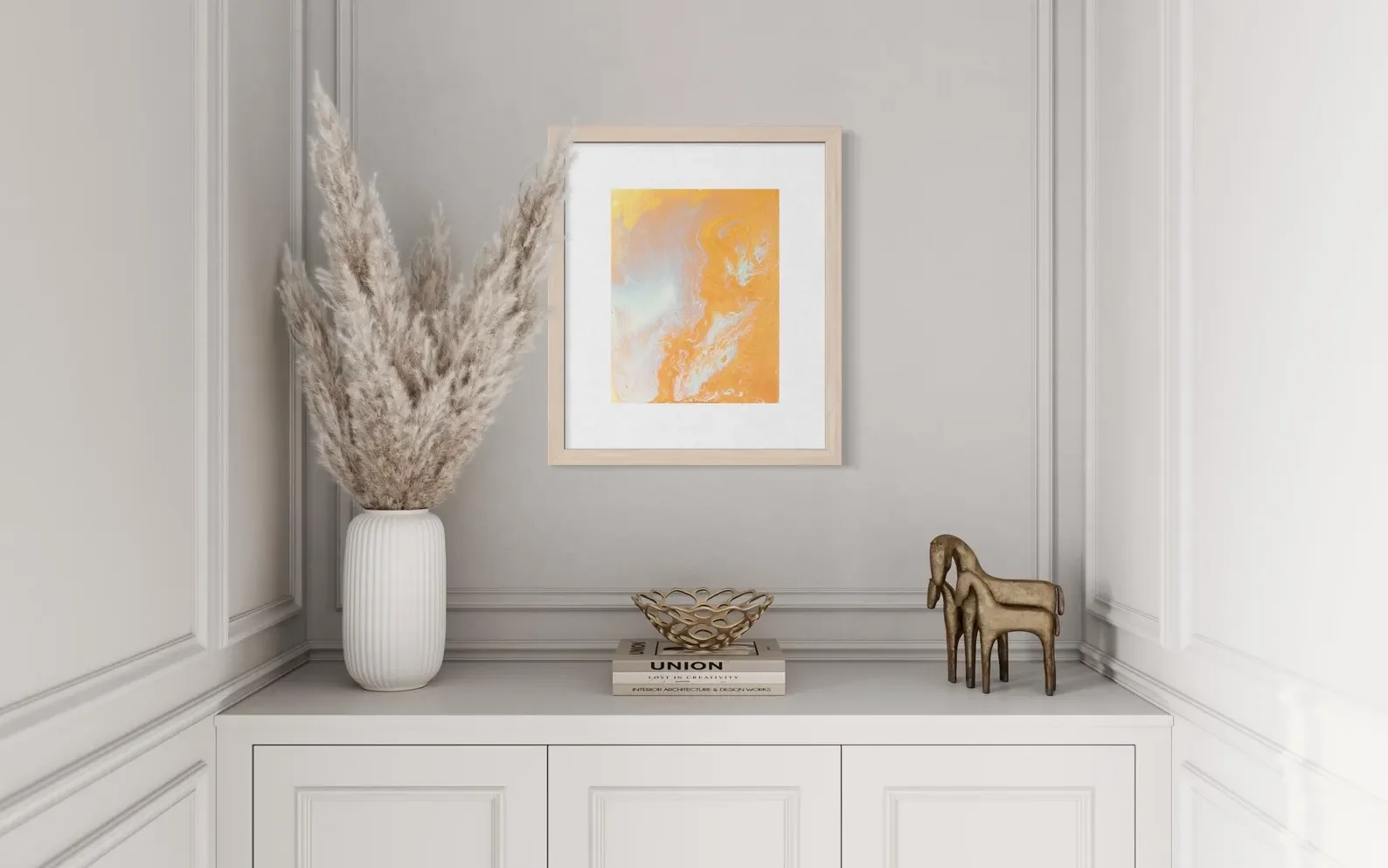 Decorative corner with a large white vase containing dried pampas grass, a framed abstract orange and pastel painting on the wall, a decorative wire bowl on top of a small stack of books, and two small gold animal figurines, all on a white sideboard.