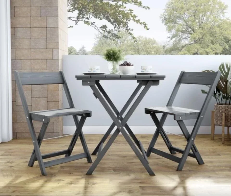 A small outdoor patio scene with a gray folding table and two matching folding chairs, a potted plant, and a frosted glass privacy screen with trees in the background.
