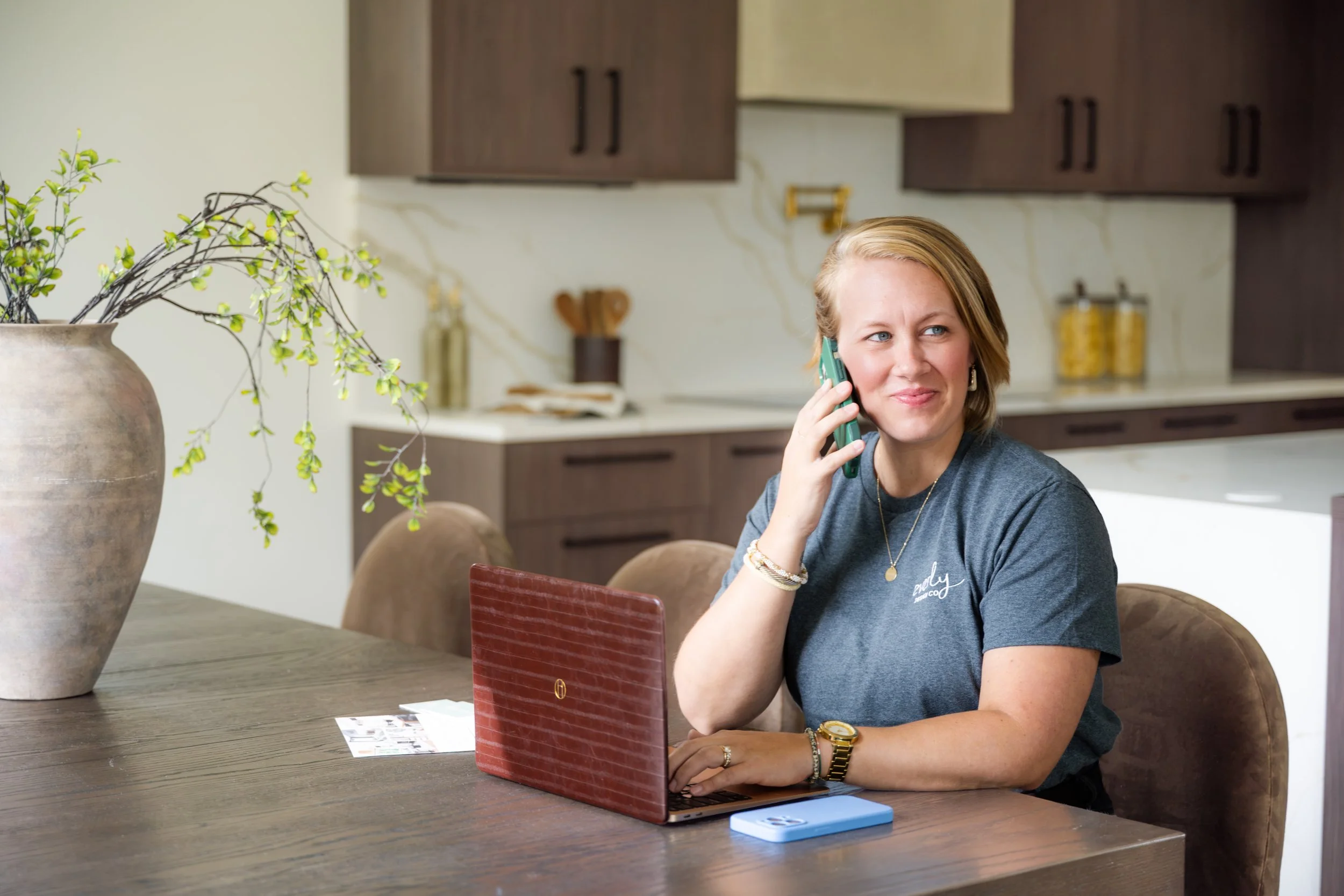 A woman with blonde hair, wearing a gray t-shirt and jewelry, is sitting at a wooden table, talking on a green phone, with a red laptop or planner in front of her, in a modern kitchen.