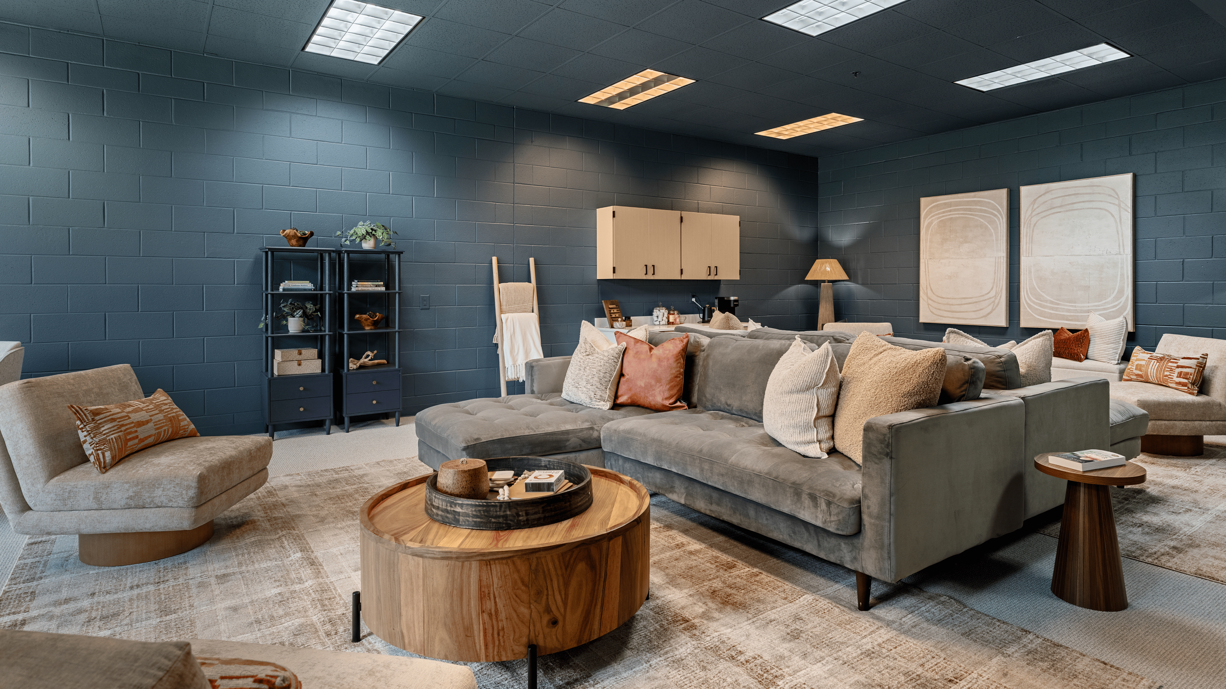 A cozy lounge area with a large gray sectional sofa, an armchair, wooden coffee tables, and decorative pillows in a room with blue brick walls and abstract artwork.
