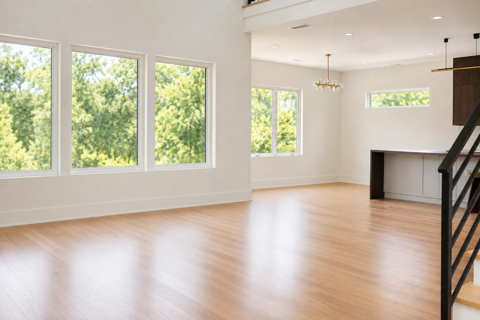 Empty living room with wooden flooring, white walls, and large windows showing green trees outside.