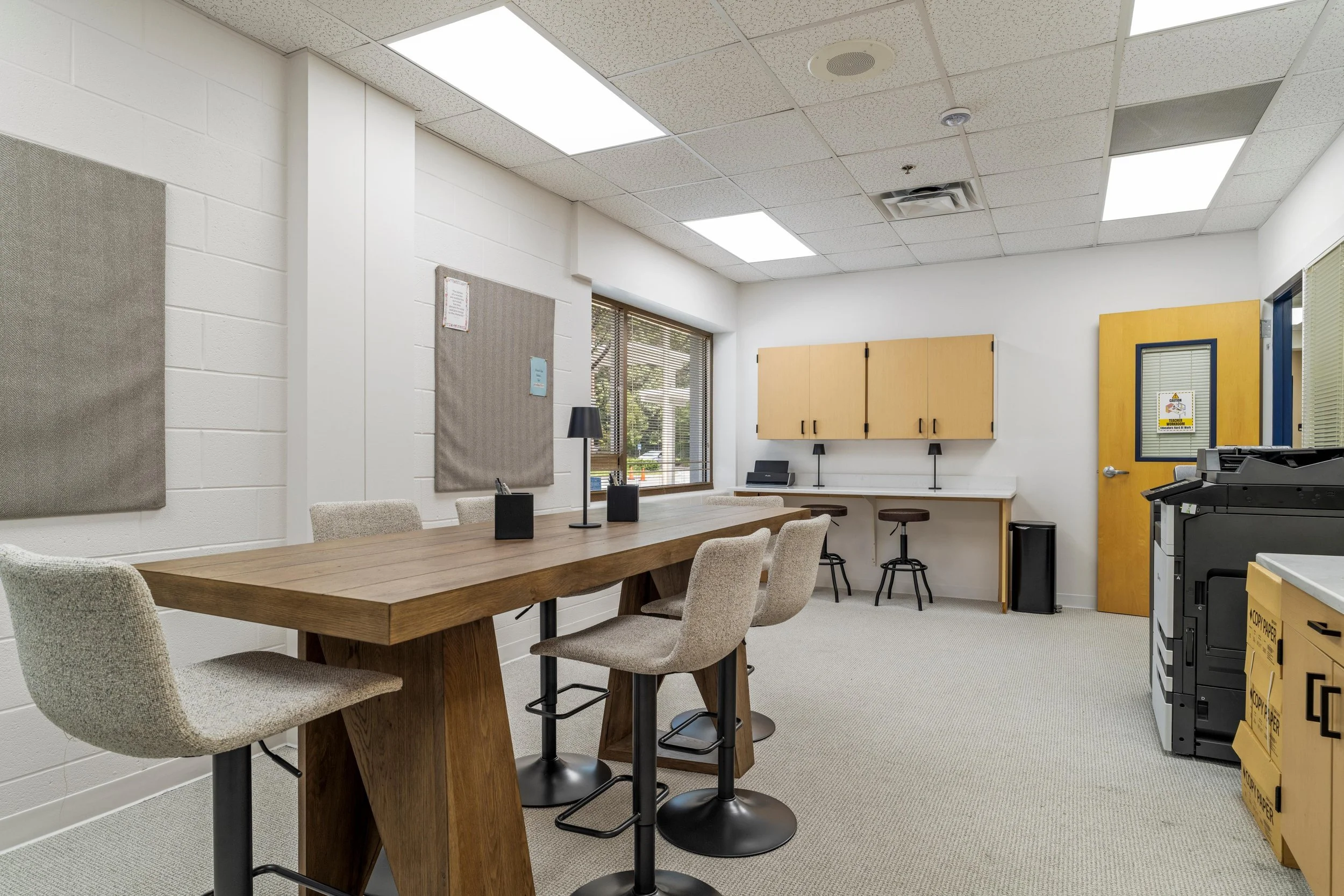 Empty classroom or meeting room with a wooden table, six beige upholstered chairs, white brick walls, grey bulletin boards, desks, cabinets, and a window with blinds.