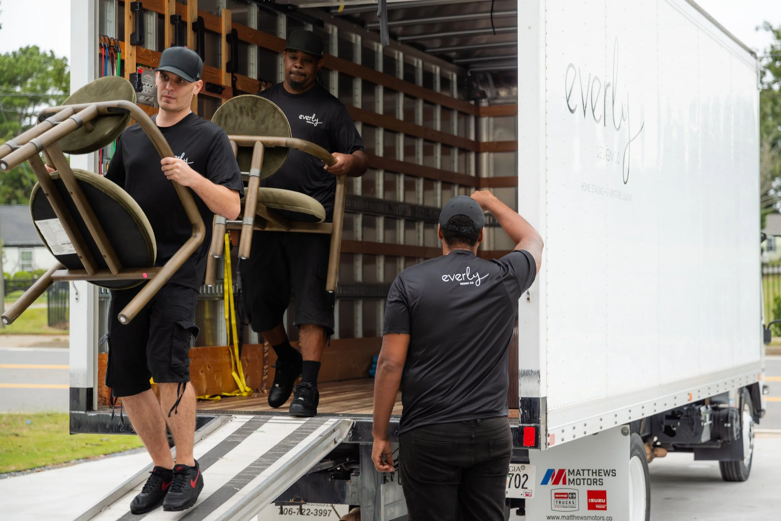 Three men are loading chairs into a moving truck. The man in front is carrying a chair, the man at the back is holding a chair, and the third man is writing on the truck wall. All are wearing black shirts with 'everly' printed on them, and the scene appears to be outdoors during daytime.