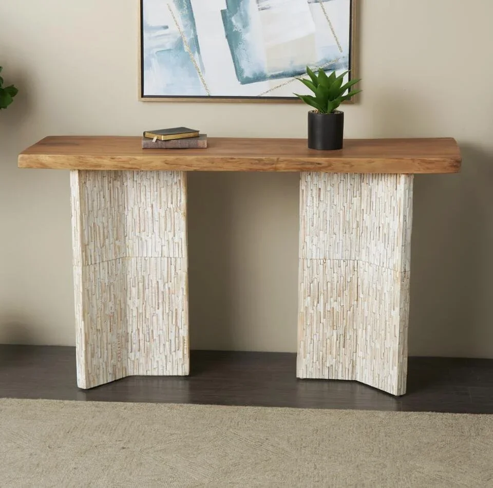 A modern console table with a natural wood top and textured beige stone or concrete legs, positioned against a wall with a painting and a small potted plant on it.