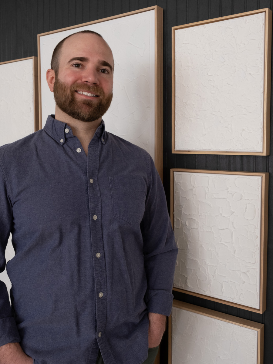 A man with a beard in a blue shirt standing in front of white textured art
