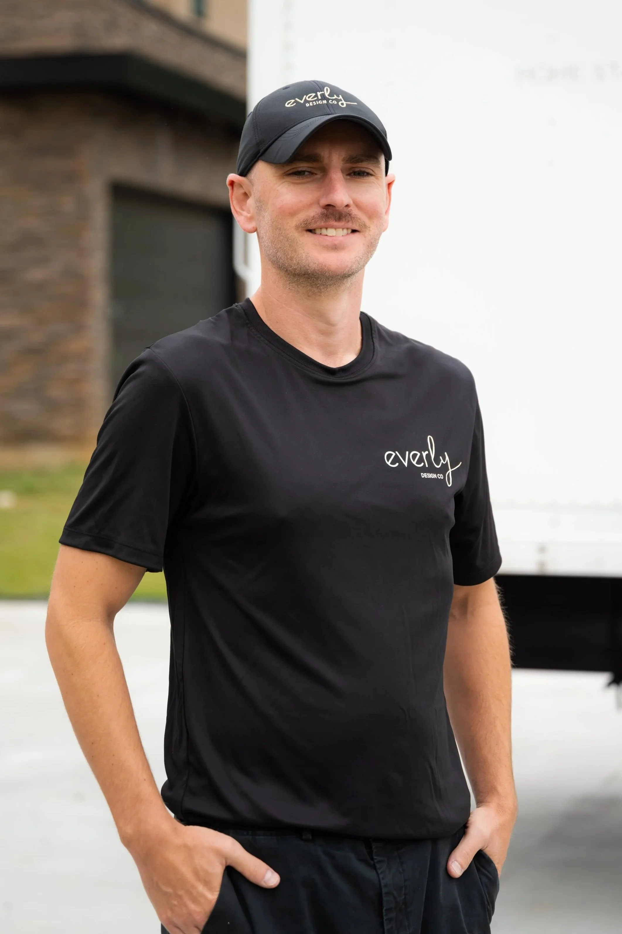 A man wearing a black cap and black t-shirt standing outdoors with his hands in his pockets, smiling at the camera, with a building and a white trailer in the background.