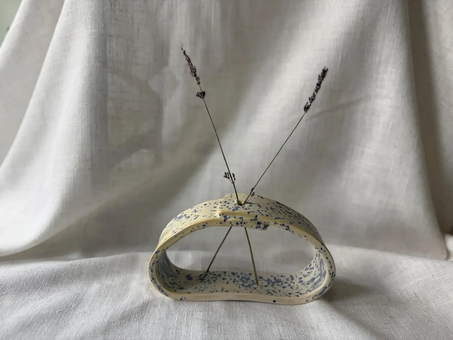 Decorative display featuring a curved ceramic sculpture with black speckles, adorned with two thin, dark purple lavender stems placed vertically, set against a light-colored fabric background.